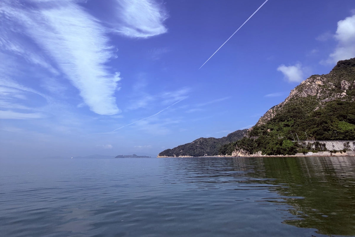 A scenic landscape photograph of a calm sea under a bright blue sky, featuring a rocky green hill on the right and wispy white clouds with contrails.